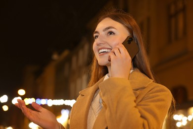 Photo of Smiling woman talking by smartphone on night city street. Space for text