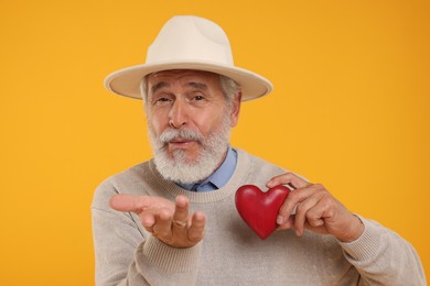 Photo of Senior man with red decorative heart blowing kiss on yellow background