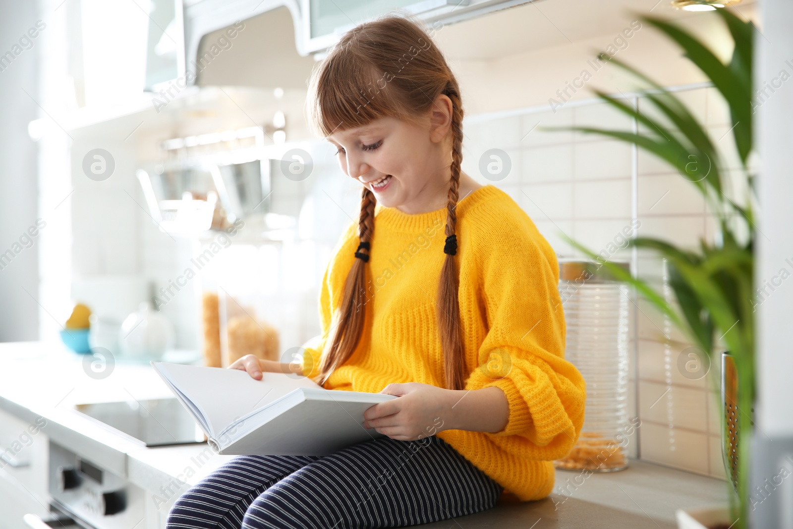 Photo of Cute little girl reading book in kitchen at home