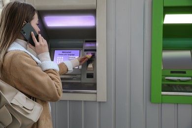 Image of Young woman talking by mobile phone while using cash machine for money withdrawal outdoors