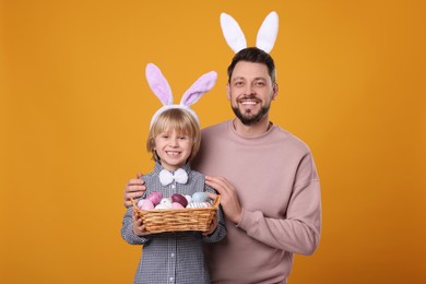 Photo of Father and son in bunny ears headbands with wicker basket of painted Easter eggs on orange background
