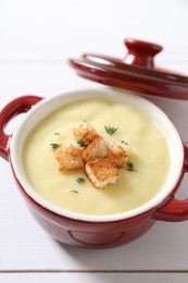 Tasty potato soup with croutons and rosemary in ceramic pot on white wooden table