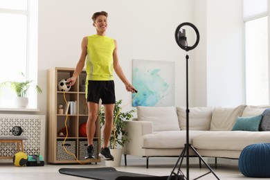 Photo of Smiling sports blogger jumping with rope while streaming online fitness lesson with smartphone at home
