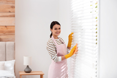 Young chambermaid wiping dust from blinds in room