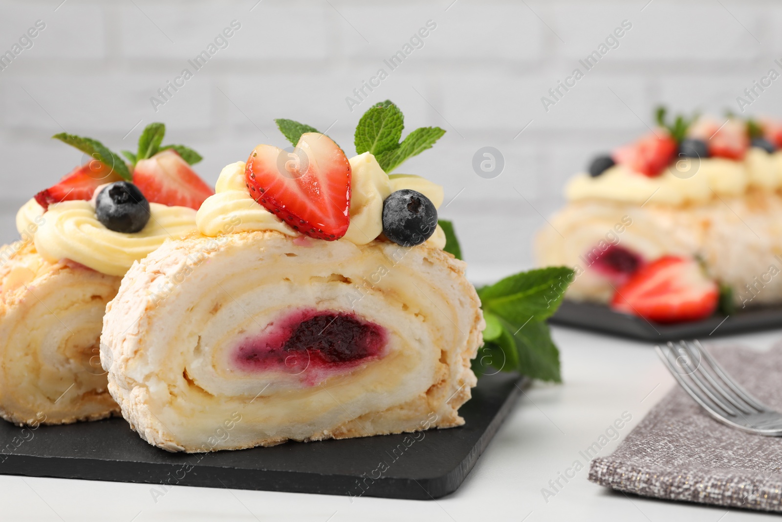 Photo of Pieces of tasty meringue roll with jam, cream, strawberry, blueberry and mint on white table, closeup. Space for text