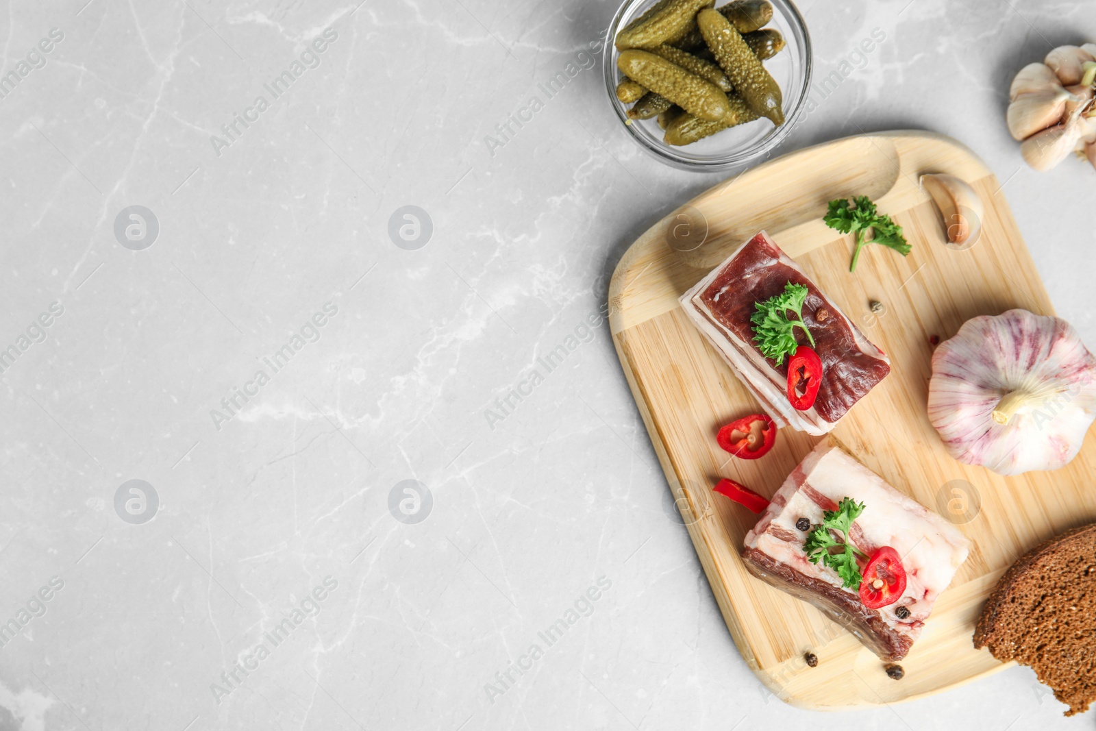 Photo of Board with pork fatback, rye bread, garlic and bowl of pickles on stone background, flat lay. Space for text