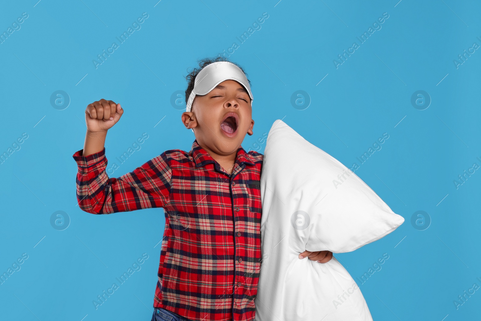 Photo of Boy with pillow and sleep mask yawning and stretching on light blue background. Insomnia problem