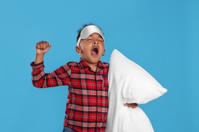 Photo of Boy with pillow and sleep mask yawning and stretching on light blue background. Insomnia problem