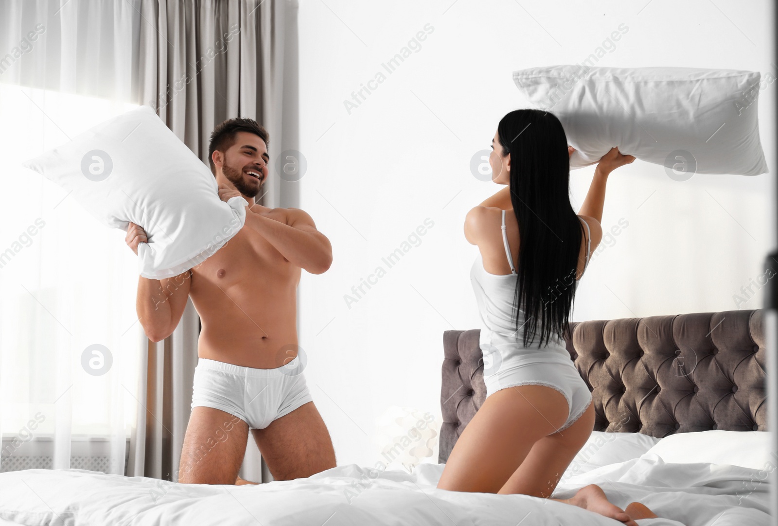 Photo of Young couple in white underwear having pillow fight on bed at home