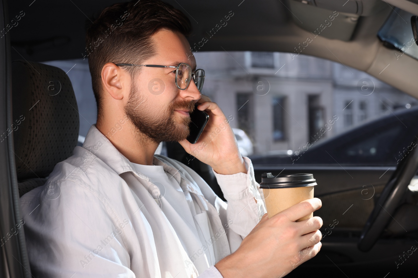 Photo of Coffee to go. Handsome man with paper cup of drink talking on smartphone in car