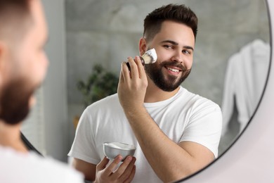 Handsome young man shaving beard near mirror in bathroom