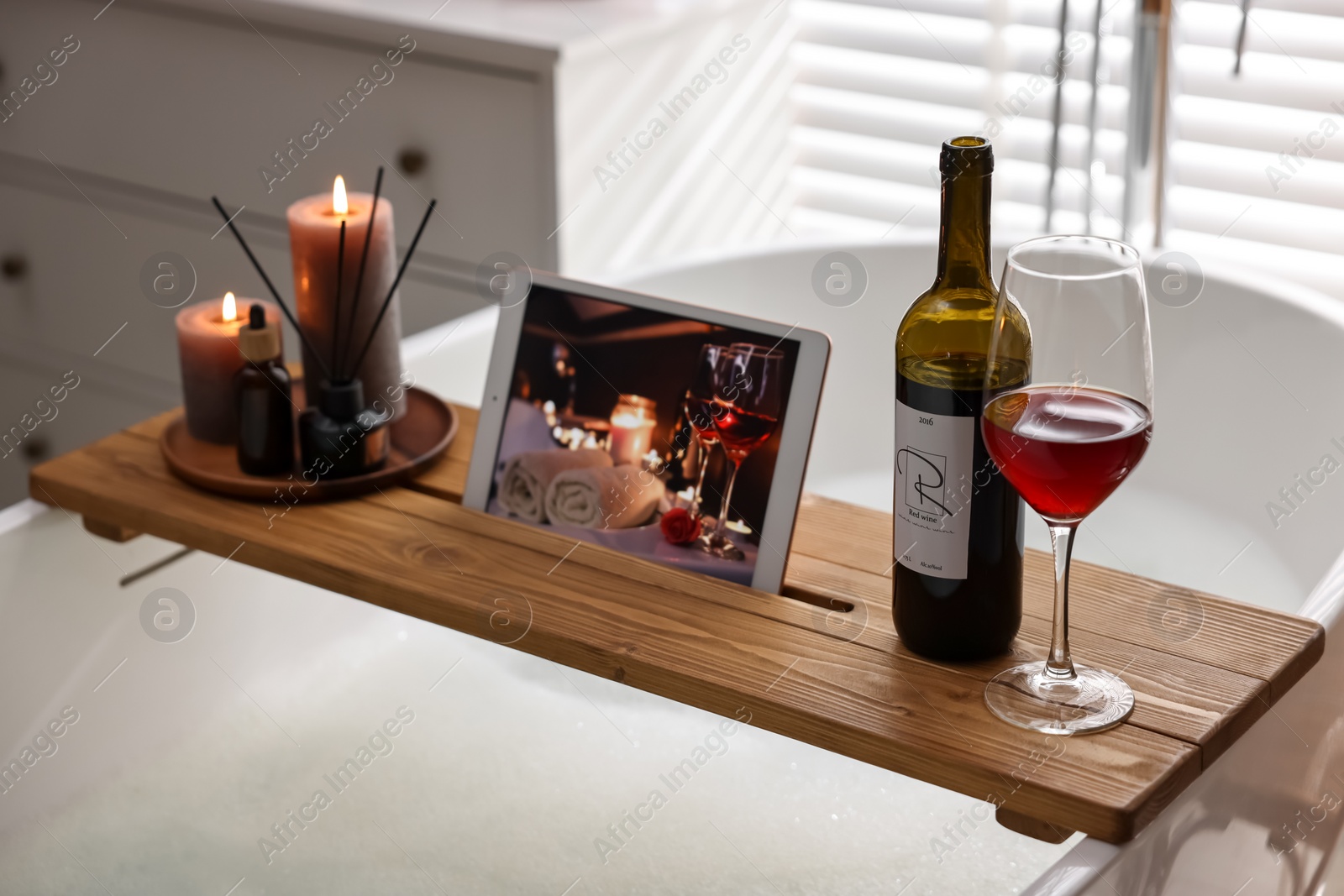 Photo of Wooden tray with tablet, wine and burning candles on bathtub in bathroom