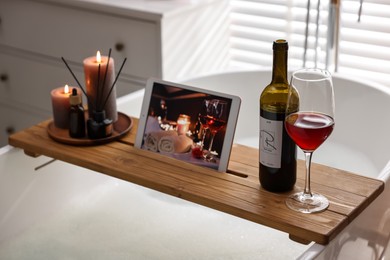 Wooden tray with tablet, wine and burning candles on bathtub in bathroom