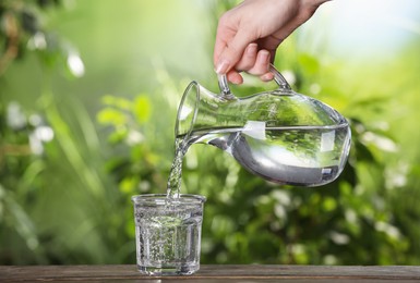 Woman pouring water from jug into glass on wooden table outdoors, closeup