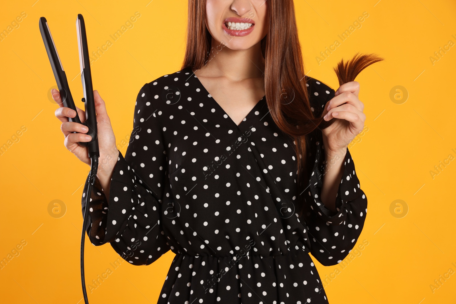 Photo of Stressed young woman with flattening iron on yellow background, closeup. Hair damage