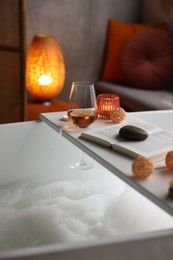 White wooden tray with glass of rose wine, book and burning candle on bathtub in bathroom