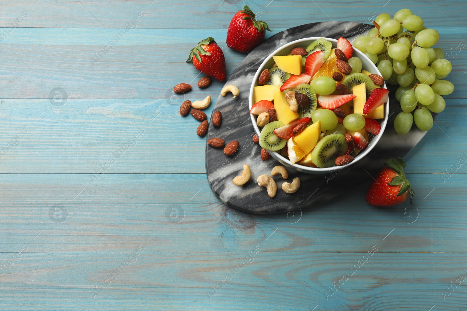 Photo of Tasty fruit salad in bowl and ingredients on light blue wooden table, flat lay. Space for text