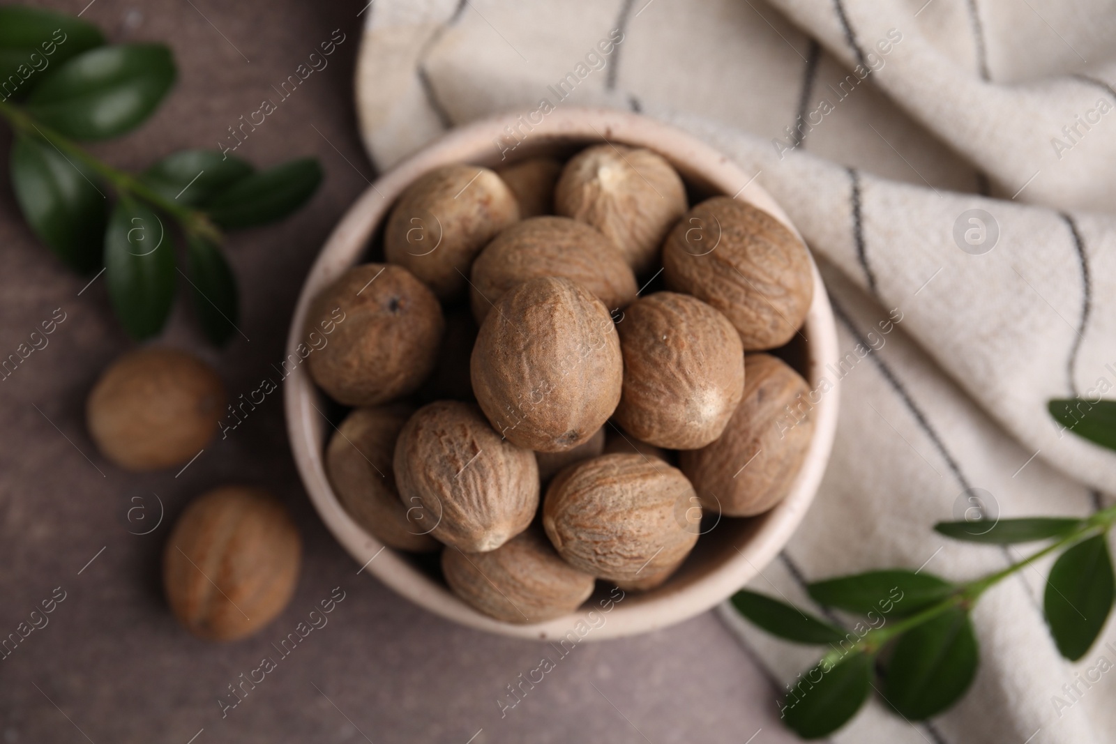 Photo of Whole nutmegs in bowl and green branches on brown table, flat lay