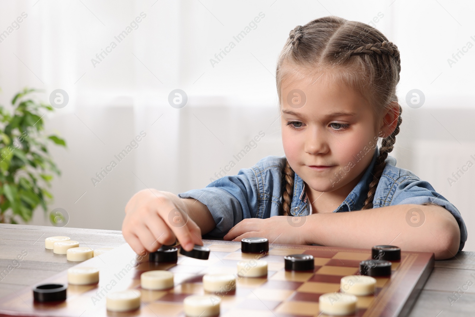 Photo of Playing checkers. Little girl thinking about next move at table in room