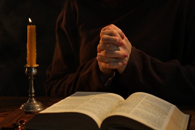 Photo of Woman praying at table with burning candle and Bible, closeup