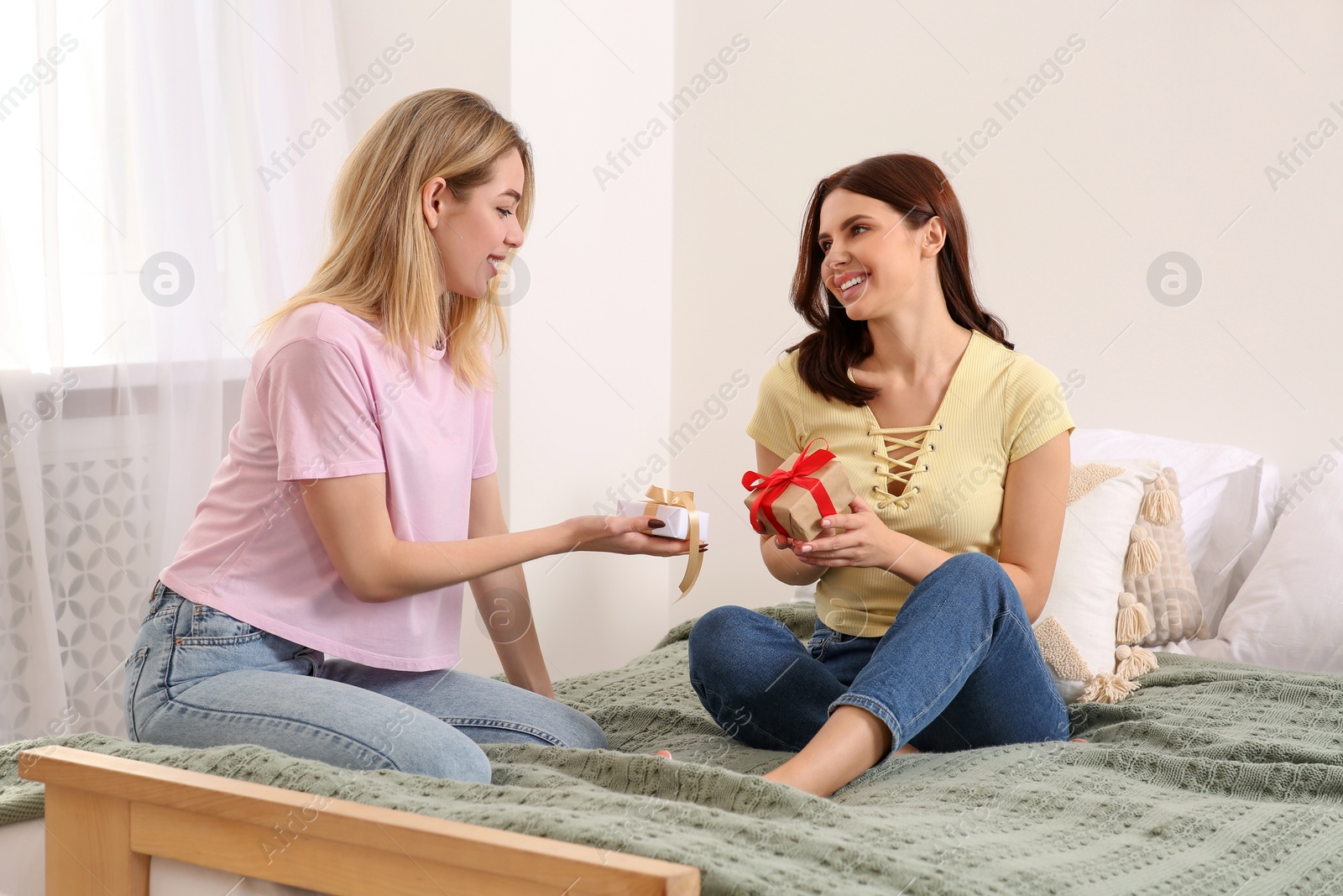 Photo of Smiling young women presenting gifts to each other on bed at home