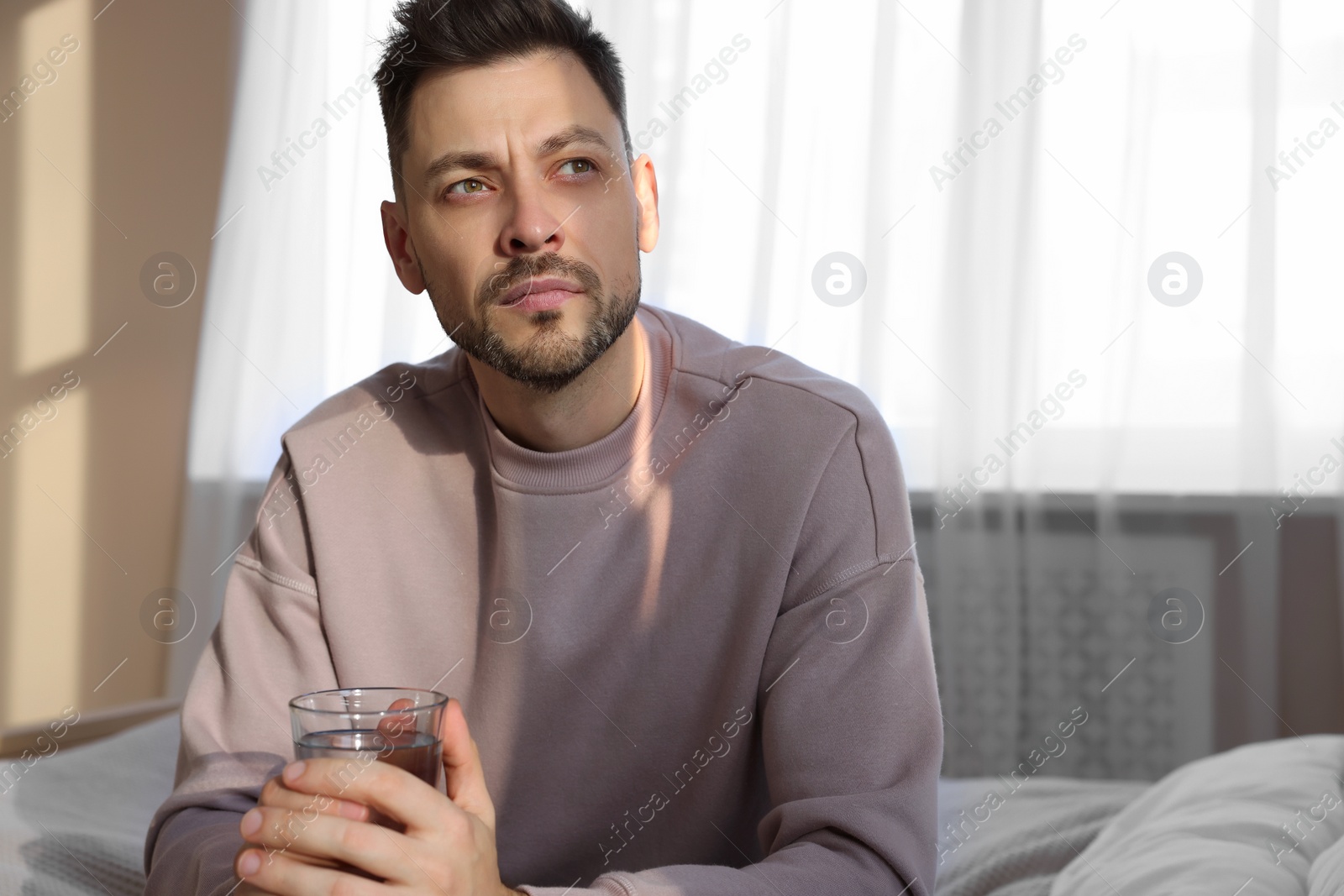 Photo of Upset man with glass of water in bedroom. Loneliness concept