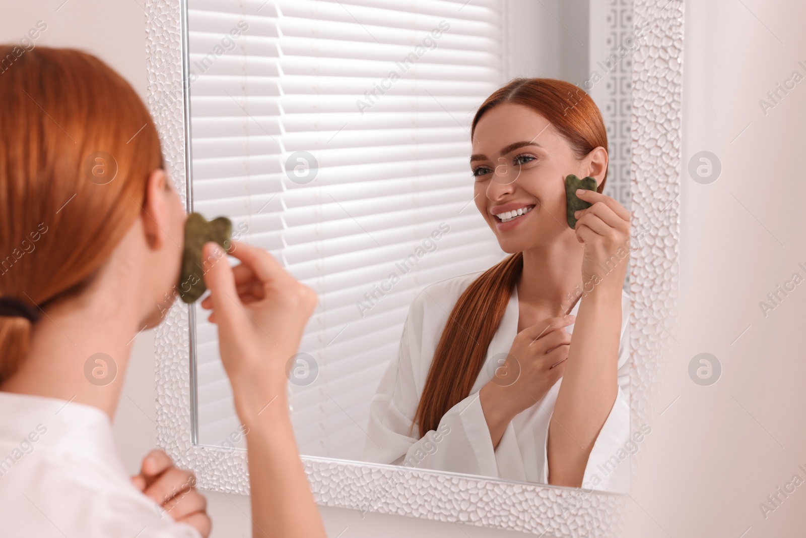 Photo of Young woman massaging her face with jade gua sha tool near mirror in bathroom