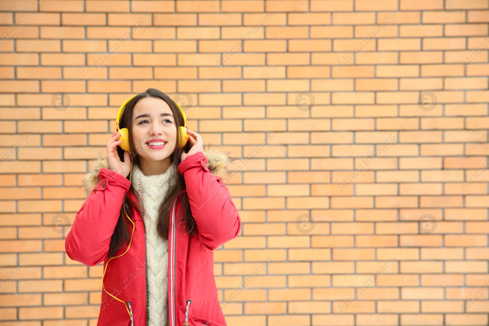 Photo of Beautiful young woman listening to music with headphones against brick wall. Space for text