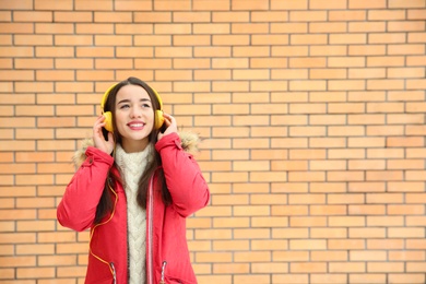Photo of Beautiful young woman listening to music with headphones against brick wall. Space for text