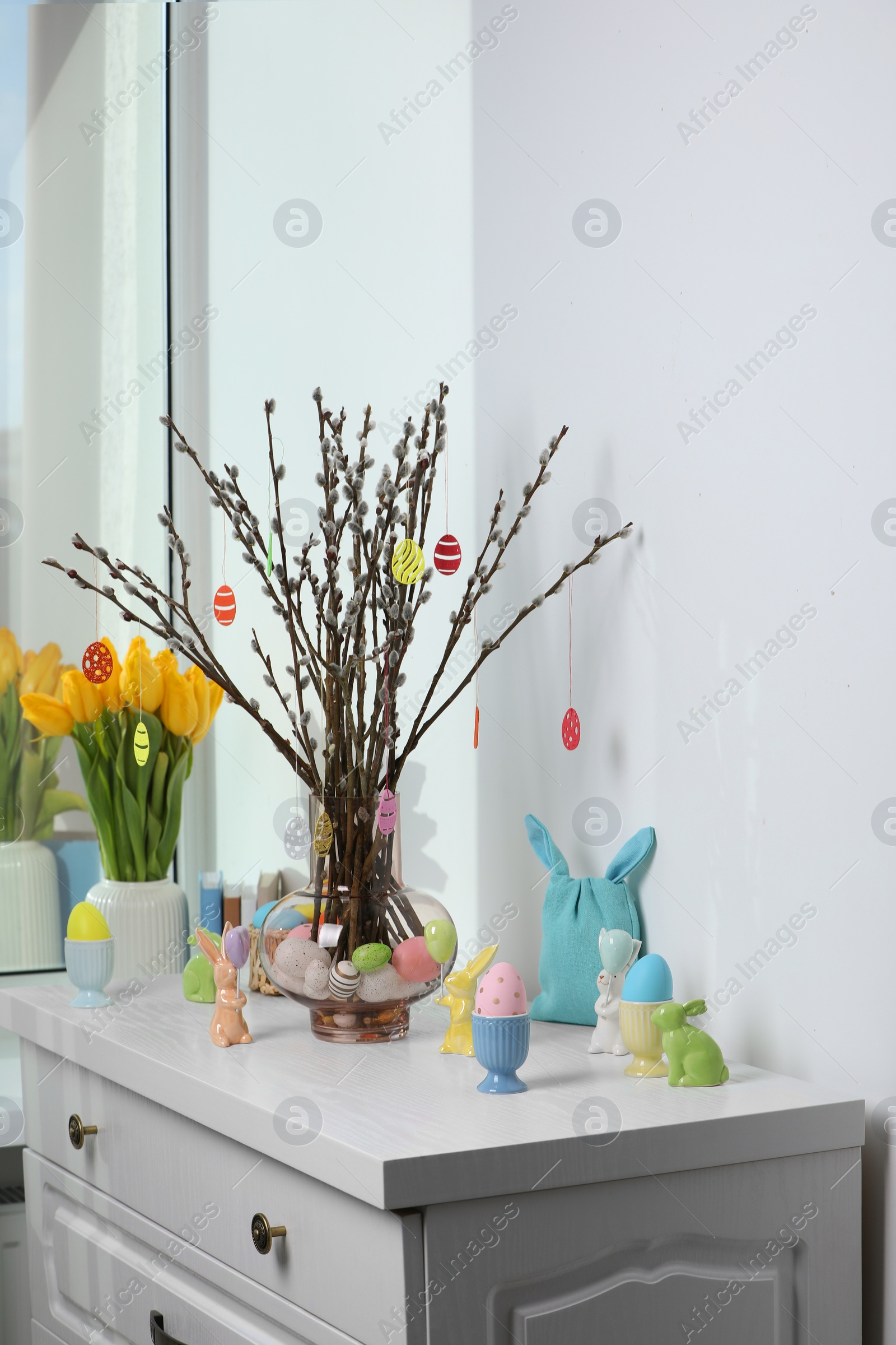 Photo of Beautiful pussy willow branches with paper eggs in vase and Easter decor on white chest of drawers at home