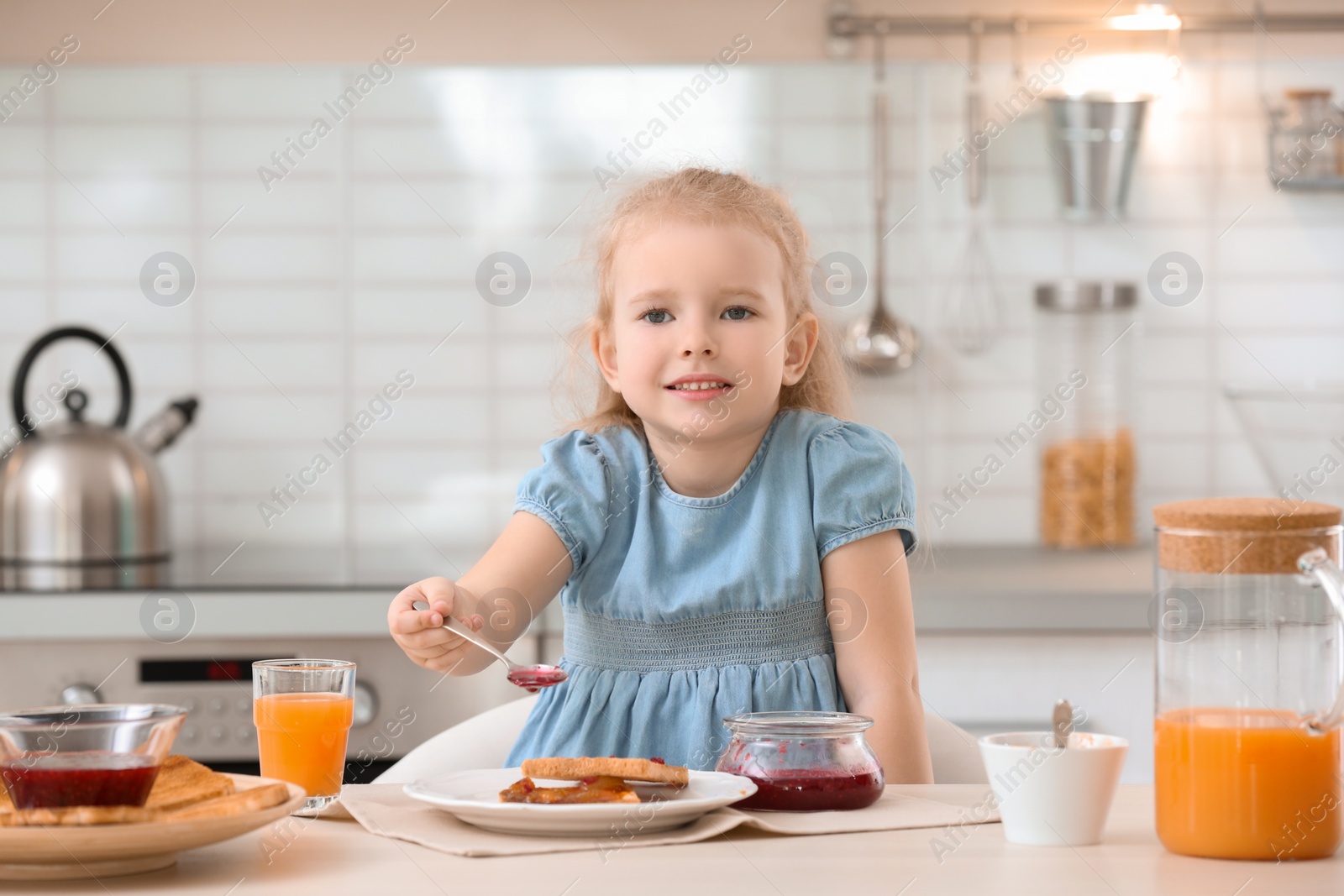 Photo of Cute little girl spreading jam onto tasty toasted bread at table in kitchen