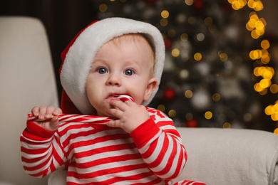 Cute baby in Santa hat and bright Christmas pajamas eating candy cane at home