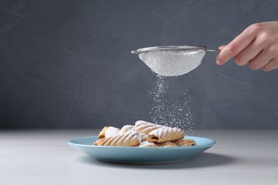 Woman with sieve sprinkling powdered sugar onto cookies at white wooden table, closeup