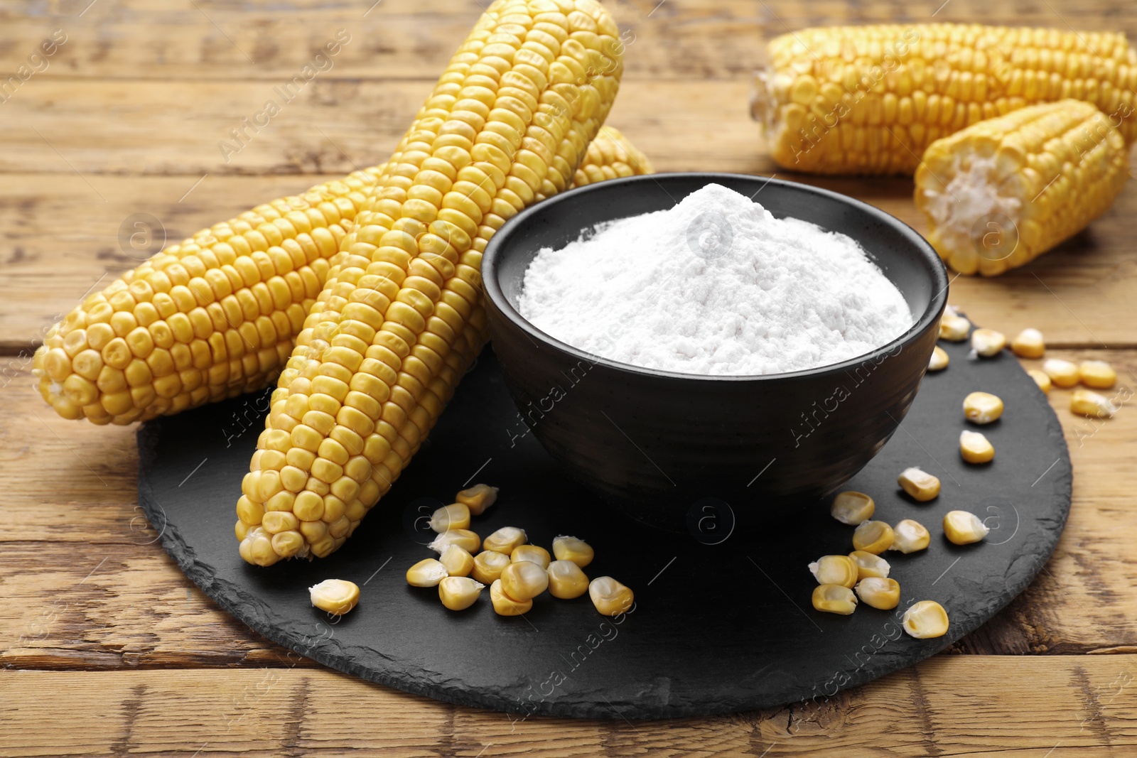 Photo of Bowl with corn starch, ripe cobs and kernels on wooden table