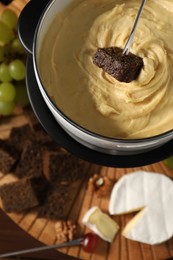 Dipping piece of bread into fondue pot with melted cheese on table with snacks, flat lay