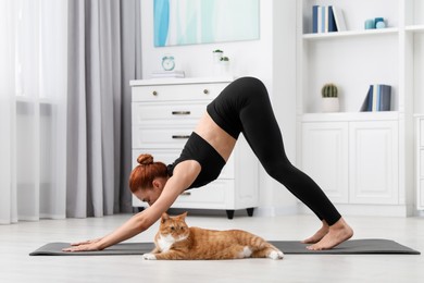 Young woman with cute red cat practicing yoga on mat at home