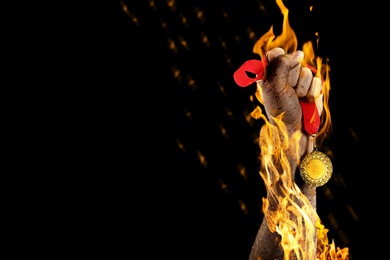Winner raising hand with gold medal from fire flames on black background, closeup. Space for text 
