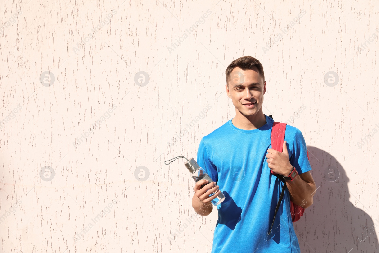 Photo of Young sporty man with backpack holding bottle of water near wall outdoors on sunny day. Space for text