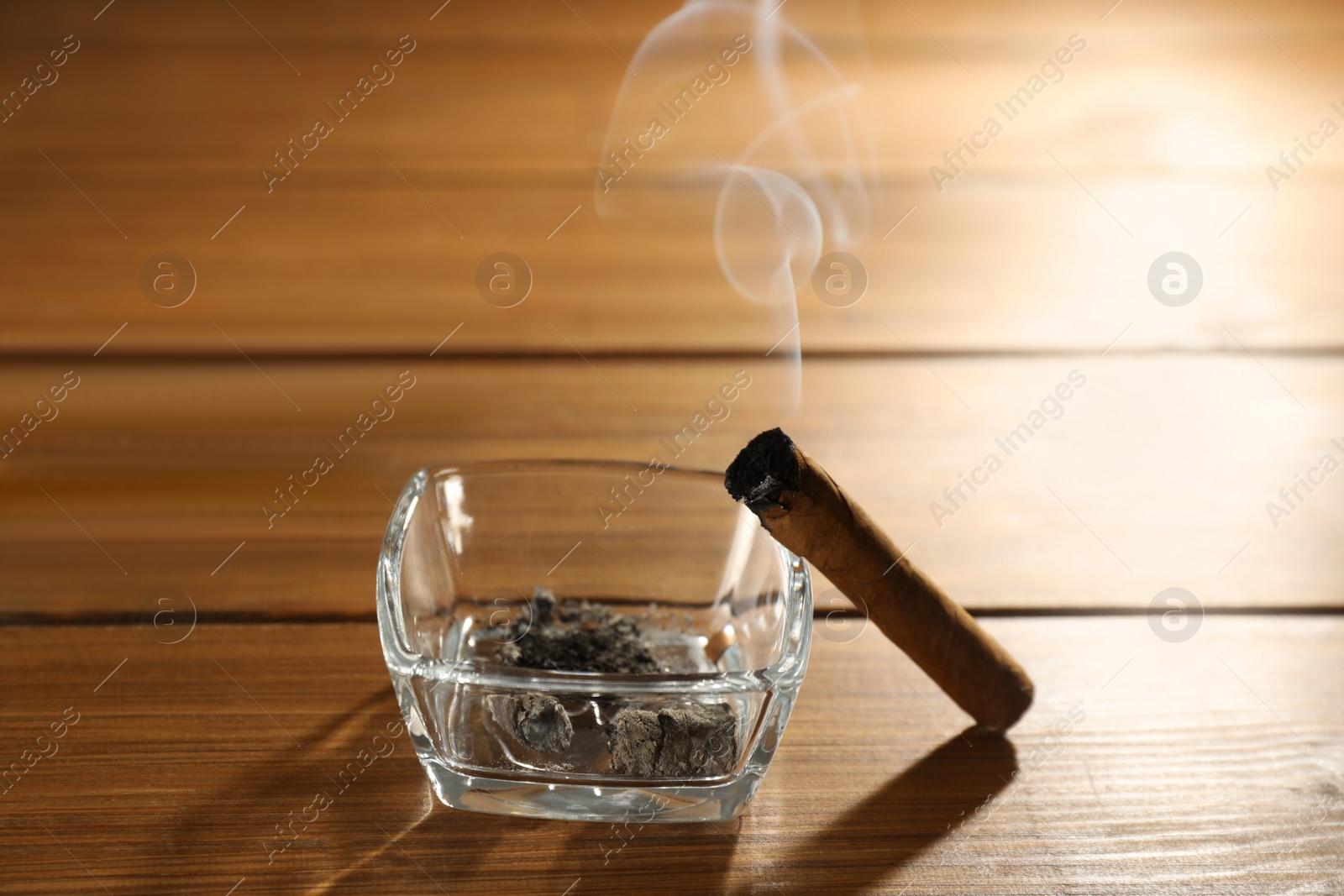 Photo of Smoldering cigar near glass ashtray on wooden table