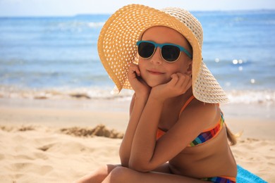 Little girl in stylish sunglasses and hat on sandy beach near sea