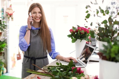 Young florist working with laptop and talking on mobile phone in flower shop