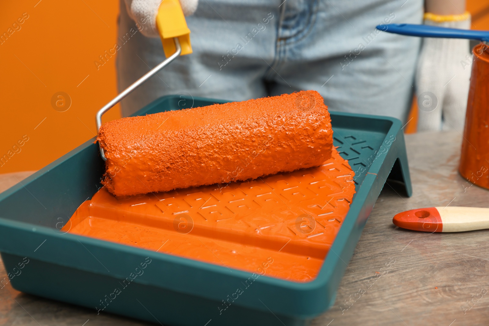 Photo of Woman taking orange paint with roller from tray at table, closeup