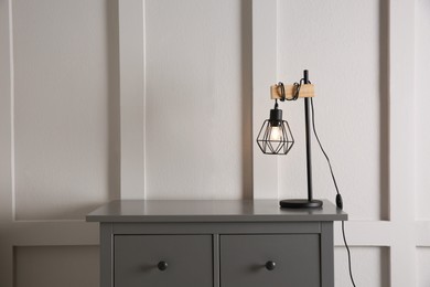 Photo of Wooden chest of drawers with modern lamp near white wall, space for text. Interior element