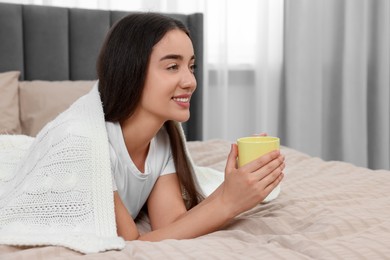 Photo of Happy young woman under plaid holding yellow ceramic mug on bed at home