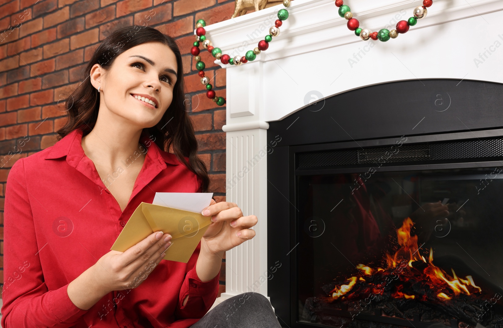 Photo of Young woman with greeting card sitting near fireplace indoors