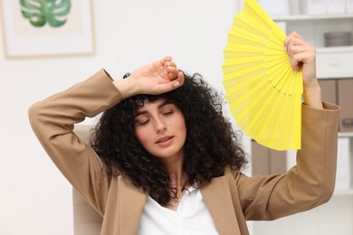 Young businesswoman waving yellow hand fan to cool herself in office