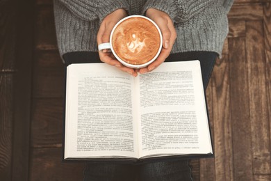 Woman with cup of coffee reading book at home, top view