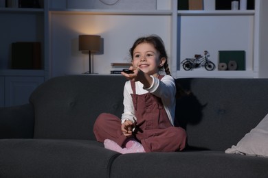 Photo of Happy girl changing TV channels with remote control on sofa at home