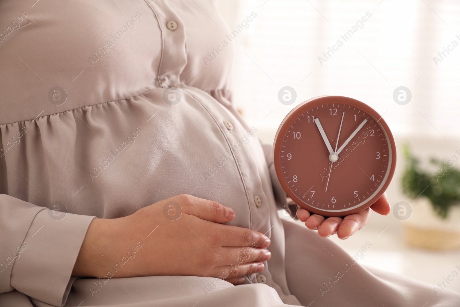 Photo of Young pregnant woman holding clock near her belly at home, closeup. Time to give birth