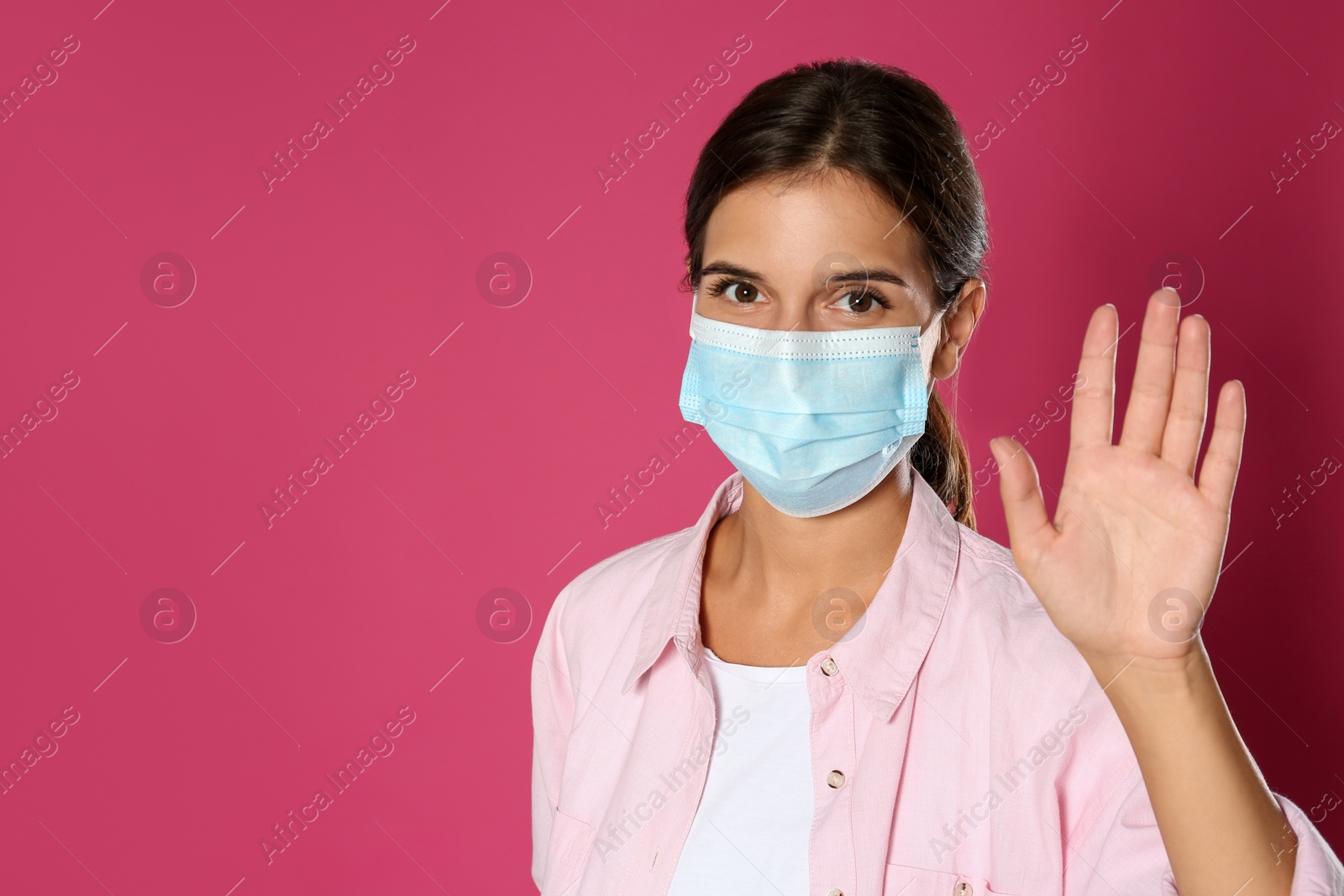 Photo of Woman in protective mask showing hello gesture on pink background, space for text. Keeping social distance during coronavirus pandemic